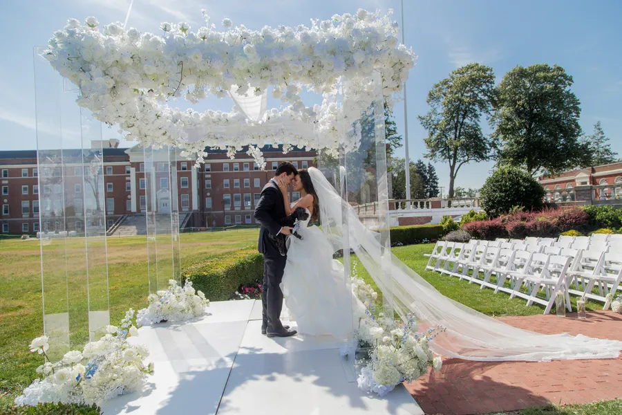 Newlyweds kissing beneath a white floral ceremony arch